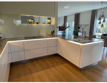 Moderne Küche mit glänzenden weißen Einbauschränken und einer Arbeitsplatte aus dunkelgrauem Material, beleuchtet durch integrierte LED-Leisten. Im Hintergrund befinden sich ein Essbereich mit Tisch und Stühlen sowie bodentiefe Fenster mit Vorhängen, die Blick auf einen Garten ermöglichen.