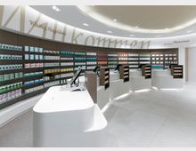 Moderner Verkaufsraum einer Apotheke mit geschwungenen weißen Tresen, mehreren Kassenstationen und Regalen an den Wänden, die mit Medikamenten- und Produktpackungen bestückt sind. Der Raum ist hell beleuchtet und an der Decke sind Begrüßungsworte in verschiedenen Sprachen angebracht.