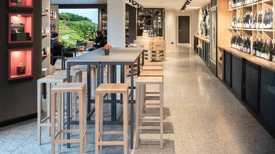 Moderner Weinladen mit langer Regalanlage aus Holz an der rechten Wand, gefüllt mit Weinflaschen, und einer zentralen hohen Tischreihe mit Holz-Hockern. Helle Innenraumbeleuchtung mit Schienenstrahlern, polierter Steinboden und ein Sitzbereich am Fenster mit Blick auf grüne Weinberge.