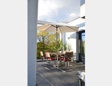 Moderne Dachterrasse mit grauem Fliesenboden, Holztisch und Stühlen mit roten Polstern unter einem großen weißen Sonnenschirm; im Hintergrund Geländer aus Glas und Bäume.