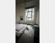 Schlichtes Schlafzimmer mit großem Sprossenfenster und weißen Vorhängen, Blick in einen Innenhof zwischen mehrstöckigen Gebäuden. Links ein niedriges Bett, auf dem Boden ein helles Kuhfell, rechts ein kleiner Schreibtisch mit transparentem Stuhl und Kommode.