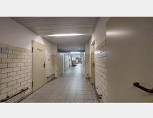 Ein langer, schlichter Flur in einem Gebäude mit hellen Fliesenwänden, grauem Bodenbelag und einer abgehängten Decke. Entlang der Wände befinden sich Türen, Handläufe und Warnhinweise; am Ende des Flurs sind metallene Transportwagen erkennbar.