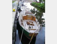 Segelboot mit Holzausstattung liegt an einer Anlegestelle in einem schmalen Kanal. Im Hintergrund sind weitere Boote und dicht bewachsene Uferböschungen zu sehen.
