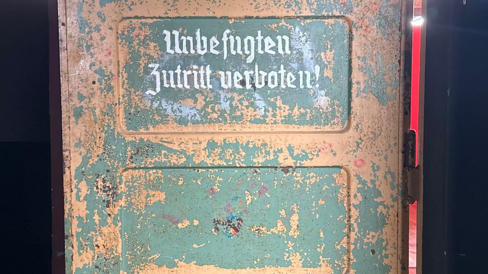 Alte, abgenutzte Holz- oder Metalltür mit abblätternder grün-beiger Farbe und mehreren eingelassenen Feldern, beschriftet mit „Unbefugten Zutritt verboten!“. Die Tür steht in einem dunklen Innenraum, seitlich fällt Licht durch eine Öffnung.