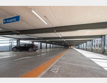 Parkebene eines modernen Parkhauses mit Betonboden, orangefarbenen Markierungen und Metallträgern. Ein schwarzes Auto ist links geparkt, und ein blaues Schild mit der Aufschrift „Ausgang“ zeigt nach links; durch die seitlichen Geländer fällt Tageslicht ein.