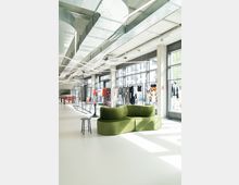 Moderner, lichtdurchfluteter Innenraum mit großen Fensterfronten, eleganten Sichtbetonelementen und freiliegenden Lüftungsschächten. Im Vordergrund steht eine grüne, geschwungene Sitzgelegenheit mit einem kleinen schwarzen Beistelltisch, während im Hintergrund Kleidung auf Ständern präsentiert wird.