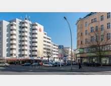 Straßenszene in einer Stadt mit einem modernen, mehrstöckigen Wohngebäude auf der linken Seite und einem historischen, beigefarbenen Gebäude mit großen Fenstern auf der rechten Seite. Im Vordergrund befinden sich eine U-Bahn-Haltestelle mit Schildern, geparkte Autos, Fahrräder und Fußgänger.