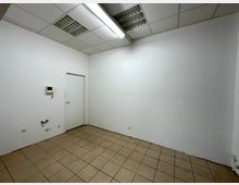 Leerer Innenraum mit weißen Wänden, braunen Fliesen auf dem Boden und einer weißen Decke mit eingelassenen Rasterleuchten. An der Wand befinden sich ein Türrahmen, Steckdosen sowie eine kleine Gegensprechanlage.