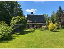 Holzhaus mit steilem Reetdach und verglaster Veranda in einem großen, gepflegten Garten mit Rasenfläche, Sträuchern und hohen Bäumen. Im Vordergrund steht eine überdachte Gartenschaukel vor dichter Bepflanzung unter blauem Himmel.