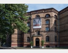Backstein-Gebäude mit historischer Architektur, gekennzeichnet durch Rundbogenfenster und Zierelemente aus Ziegeln. Über dem Eingang hängt ein großes Banner mit der Aufschrift "Eventlocation", flankiert von zwei Pflanzentöpfen, und im Vordergrund stehen Bäume.