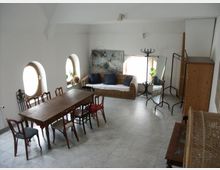 Ein heller, geräumiger Wohnbereich mit weiß gestrichenen Wänden, runden Fenstern und Fliesenboden. Im Vordergrund steht ein langer Holztisch mit unterschiedlichen Stühlen, im Hintergrund eine Sitzgruppe mit einem Sofa aus Rattan, Spiegel und Kleiderständer.