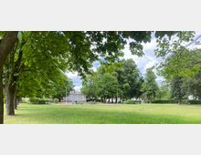 Grüne Parklandschaft mit Rasenfläche und dichtem Baumbestand, die durch überhängende Äste eingerahmt wird. Im Hintergrund ist ein steinernes Denkmal sowie eine kleine gepflasterte Fläche zu erkennen.