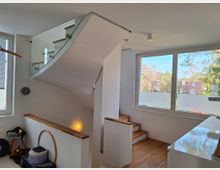 Innenraumansicht mit einer modernen Holztreppe, die mit einem Glasgeländer ausgestattet ist. Große Fenster lassen viel Tageslicht herein, und im Hintergrund sind Bäume sowie Hausdächer zu sehen.