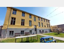 Gelbes, dreistöckiges Gebäude mit großen rechteckigen Fenstern und grauem Sockelbereich, davor ein grüner Rasen mit Gartenbänken, Tischen und einem blauen Holz-Picknicktisch. Im Hintergrund sind eine gepflasterte Terrasse und ein Nebengebäude aus Holz und Ziegeln zu sehen.