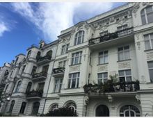 Mehrstöckiges historisches Wohngebäude im Stil des Spätklassizismus, mit reich verzierten weißen Fassaden, großen Fenstern und Balkonen mit schmiedeeisernen Geländern. Vor dem Gebäude ist ein kleiner Gartenbereich zu sehen, der Himmel ist blau mit wenigen Wolken.