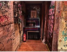 Schmaler Innenflur mit Holzboden, dessen Wände und Türen dicht mit Graffiti und Aufklebern bedeckt sind. Am Ende des Gangs stehen ein beleuchteter Automat bzw. eine Jukebox mit Tastaturfeld sowie ein Feuerlöscher und eine niedrige Box auf dem Boden.