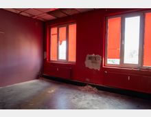 Leerer Innenraum mit dunkelroten Wänden und zwei großen Fenstern, die mit roten Rollos abgedeckt sind; unter den Fenstern verlaufen Heizkörper. An der Wand sind beschädigte Putzstellen und Bauschutt am Boden sichtbar, die Decke besteht aus Deckenplatten.