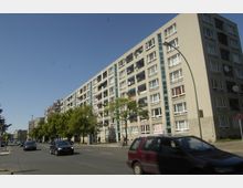 Mehrstöckiges Wohngebäude in Plattenbauweise an einer befahrenen Straße mit geparkten Autos und einzelnen Bäumen entlang des Gehwegs. Die Fassade zeigt viele Fenster und Balkone mit Satellitenschüsseln.