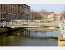 Eine Fußgängerbrücke aus Metall und Holz überquert einen ruhigen Wasserkanal, flankiert von historischen Gebäuden mit aufwendiger Fassade und Rundbogenfenstern. Im Hintergrund befinden sich weitere Gebäude mit roten Dächern sowie eine gepflasterte Fläche mit Bänken und vereinzelten Bäumen.