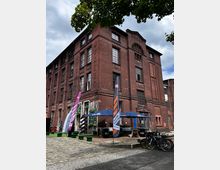 Backstein-Gebäude mit mehreren Stockwerken und großen Sprossenfenstern, im Vordergrund ein gepflasterter Platz mit Sitzgelegenheiten, Sonnenschirmen und Werbefahnen; Fahrräder stehen an einer Seite des Platzes. Der Himmel ist bewölkt, und ein Baum ragt am oberen Bildrand ins Bild.