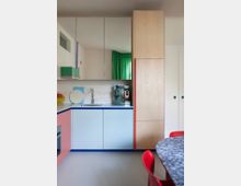 Moderne Küche mit farbenfrohem Design, bestehend aus pastellblauen und lachsfarbenen Schränken, Holzelementen, einer Spüle, einer Kaffeemaschine und Spiegelschränken. Im Vordergrund steht ein Tisch mit weiß-grau gemusterter Marmorplatte und roten Stühlen, während ein grüner Vorhang im Hintergrund zu sehen ist.