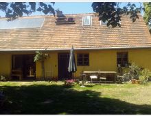 Ein gelbes Haus mit einem roten Ziegeldach und Dachfenstern, umgeben von einem Garten mit Rasenfläche. Im Vordergrund stehen ein Holztisch mit Stühlen, ein geschlossener Sonnenschirm und Blumenkübel; an der Hauswand lehnen ein Fahrrad und weitere Gegenstände.