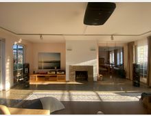 Wohnzimmer mit heller Einrichtung, einem zentralen Kamin und großen Fenstern, die den Raum mit Tageslicht durchfluten. Links und rechts des Kamins stehen schwarze Lautsprecher, und im Hintergrund ist ein Essbereich mit Holzmöbeln erkennbar.