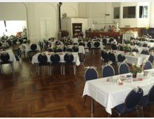 Großer Veranstaltungsraum mit Holzboden, mehreren festlich gedeckten Tischen mit weißen Tischdecken und dekorierten Stühlen. Im Hintergrund befinden sich eine Theke, ein Spiegel und hohe Fenster, die den Raum mit Tageslicht erhellen.