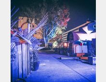 Beleuchteter Außenbereich bei Nacht mit kreativen Holzkonstruktionen und dekorativen Pflanzenkübeln. Im Hintergrund sind vereinzelte Bäume sowie ein modernes Mehrstöckengebäude zu sehen.