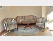 Sitzbereich mit einer Holzcouch und zwei passenden Sesseln mit gestreiftem Stoffbezug, einem ovalen Holztisch in der Mitte und einem ovalen Teppich auf einem Holzboden. Die Wände sind cremefarben, und der Raum ist von Sonnenlicht durchflutet.