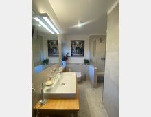 Modernes Badezimmer mit einem langen Waschbecken auf einer Holzkonsole, einer Toilettenschüssel und einer begehbaren Dusche mit Glasabtrennung. Wände und Boden sind mit hellen Fliesen verkleidet, und an der Wand hängen zwei Bilder mit tropischem Motiv.