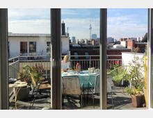 Blick durch eine Glastür auf eine Dachterrasse mit Tisch, Stühlen und Pflanzen. Im Hintergrund sind Wohngebäude und der Berliner Fernsehturm zu sehen.