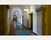 Langer Flur mit Rundbogen-Decke in einem historischen Gebäude, beleuchtet von Hängelampen. Rechts befindet sich eine Aufzugsöffnung mit einem leuchtenden "Aufzug"-Schild, links sind Fenster und Heizkörper zu sehen.