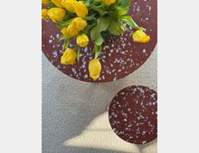 Top-down view of a round, red terrazzo coffee table with white flecks holding a glass vase of yellow tulips, set on a light textured carpet. A smaller matching terrazzo side table sits nearby, with sunlight casting a shadow across the floor.