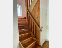 Holztreppe in einem Wohnraum mit naturbelassenen Holzstufen und einer holzvertäfelten Wand. Rechts daneben befindet sich eine weiße Zimmertür, und im Hintergrund ist ein Fenster mit Blick ins Grüne sichtbar.