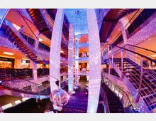 Beleuchteter Innenbereich eines modernen Gebäudes mit mehreren Etagen, Treppen und Geländern aus Metall. Eine große Discokugel hängt in der Mitte, und farbige Lichtpunkte werden auf die Wände projiziert.