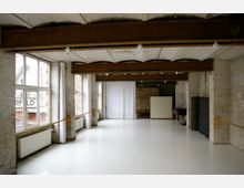 Großer Raum mit hohen Decken und sichtbaren Betonbalken, teils verputzten Ziegelwänden und großen Fenstern, die Tageslicht hereinlassen. Der Boden ist mit hellem, glänzendem Material bedeckt, an einer Seite sind Wandstangen angebracht, vermutlich für Ballett oder Tanzübungen.