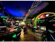 Innenraum einer Bar mit geschwungener Holztheke, hohen Tischen und Barhockern auf Holzboden, beleuchtet durch grüne und blaue Neonlichter. An den Wänden hängen Leuchtreklamen, und unter der dunklen Decke verlaufen sichtbare Metalltraversen mit Lichttechnik.