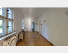 Langer, heller Flur mit weiß gestrichenen Wänden, Holzdielenboden und großen Fenstern auf der linken Seite. Rechts befinden sich Wandlampen und Türen, die zu angrenzenden Räumen führen.