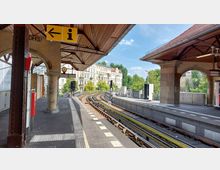 Bahnhof einer oberirdischen U-Bahn-Station mit überdachten Bahnsteigen und einem gewölbten Dach aus Holz. Im Hintergrund erstrecken sich Gleise in eine Kurve, mit angrenzenden Wohngebäuden und grünen Bäumen im Stadtumfeld.