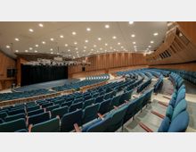 Ein moderner, großer Veranstaltungssaal mit ansteigend angeordneten, gepolsterten blauen Sitzreihen, einer breiten Bühne mit schwarzem Vorhang und Holzverkleidung an den Wänden. Die Decke ist mit zahlreichen eingebauten Leuchten ausgestattet, und über der Bühne befindet sich ein Gestell mit Scheinwerfern.