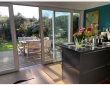 Moderne Küche mit Blick auf eine Terrasse durch große Glasschiebetüren, die viel Tageslicht hereinlassen. Im Vordergrund eine Kücheninsel mit Blumenstrauß, und draußen befinden sich Holzmöbel sowie ein gepflegter Garten mit Bäumen und Sträuchern.