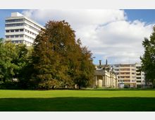 Ein Park mit einer großen Rasenfläche im Vordergrund, umgeben von Bäumen. Im Hintergrund sind ein mehrstöckiges modernes Bürogebäude, ein historisches Gebäude mit Säulen und Statuen sowie Wohnhäuser zu sehen.