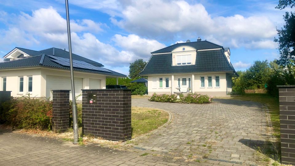 Einfahrt zu einem Wohnhaus mit heller Fassade und dunkel gedecktem Mansarddach, davor ein gepflasterter Hof und ein niedriges Blumenbeet. Links steht ein separates Garagengebäude mit Solarpaneelen auf dem Dach, am Straßenrand sind dunkle Ziegelpfeiler und eine Laterne zu sehen.