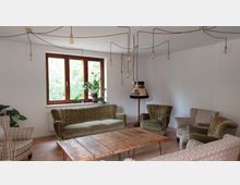 Wohnzimmer mit einer Sitzgruppe aus grünlichen und gemusterten Polstermöbeln, einem rustikalen Holztisch und einer Stehlampe mit Stoffschirm. Im Hintergrund befindet sich ein großes Fenster mit Topfpflanzen auf der Fensterbank, und von der Decke hängen mehrere freiliegende Glühbirnen an schwarzen Kabeln.