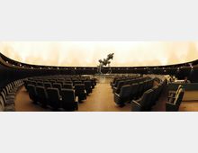 Ein Innenraum eines Planetariums mit einem zentral positionierten Projektor in der Mitte, umgeben von halbkreisförmig angeordneten Reihen gepolsterter Sitze. Die Kuppeldecke ist hell beleuchtet, und die dunklen Wände enthalten integrierte Beleuchtungselemente.