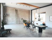 Ein modernes Loft mit minimalistischem Interieur, bestehend aus einer Betonwand, einem langen Holztisch mit bunten Stühlen, einer Leiter an der Wand und Pflanzen als Dekoration. Im Hintergrund befinden sich ein großes Fenster, eine Studiolampe und eine hängende Einrichtung mit Körben.