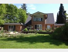 Backsteinhaus mit dunklem Satteldach und modernem Balkon, umgeben von einem gepflegten Garten mit Rasenfläche, Blumenbeeten und Bäumen im Hintergrund. Das Gebäude hat mehrere Fensterfronten, die eine helle, einladende Atmosphäre vermitteln.