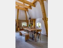 Esszimmer in einem rustikalen Dachraum mit sichtbaren Holzbalken, einem Holztisch mit Stühlen und einer Bank. Im Hintergrund befinden sich kleine Fenster mit Blumen und ein Schrank an einer gemauerten Wand.