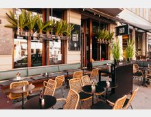 Außenbereich eines Straßencafés mit mehreren runden Tischen und Flechtstühlen sowie einer langen gepolsterten Sitzbank entlang der Fassade. Über den Fenstern hängen Pflanzkästen mit Palmen, und Markisen sowie ein Schild „Bar 1000“ rahmen den Eingangsbereich.